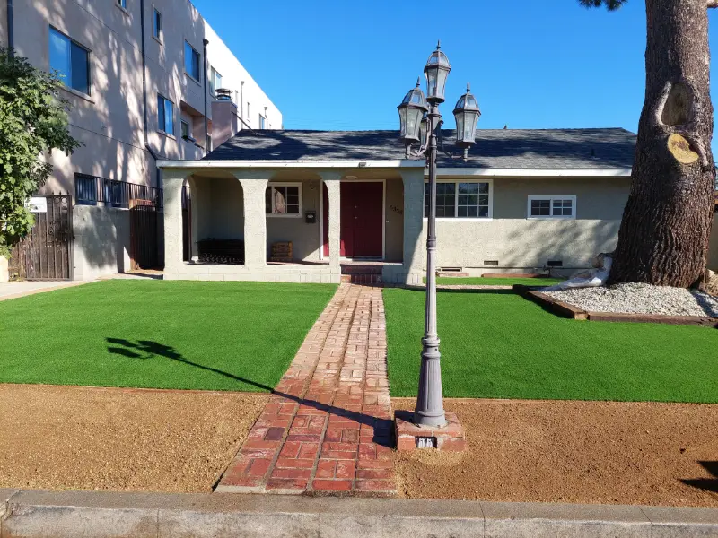 Artificial turf lawn edged by a brick paver walkway leading to a house entrance with a lamp post
