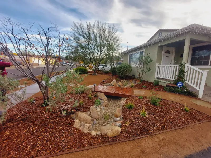 Front yard landscaped with red bark mulch, boulders and a young tree in front of a beige porched house